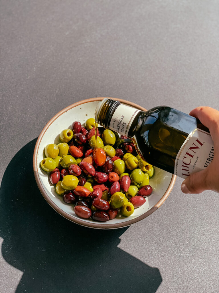 Citrus-marinated olives with herbs in a bowl with olive oil being poured over them.
