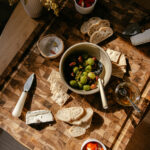 Rosemary and citrus-marinted olives in a small bowl on a wooden board surrounded by crackers, cheese, and bite-size bread.