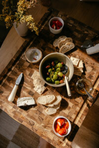 Rosemary and citrus-marinted olives in a small bowl on a wooden board surrounded by crackers, cheese, and bite-size bread.