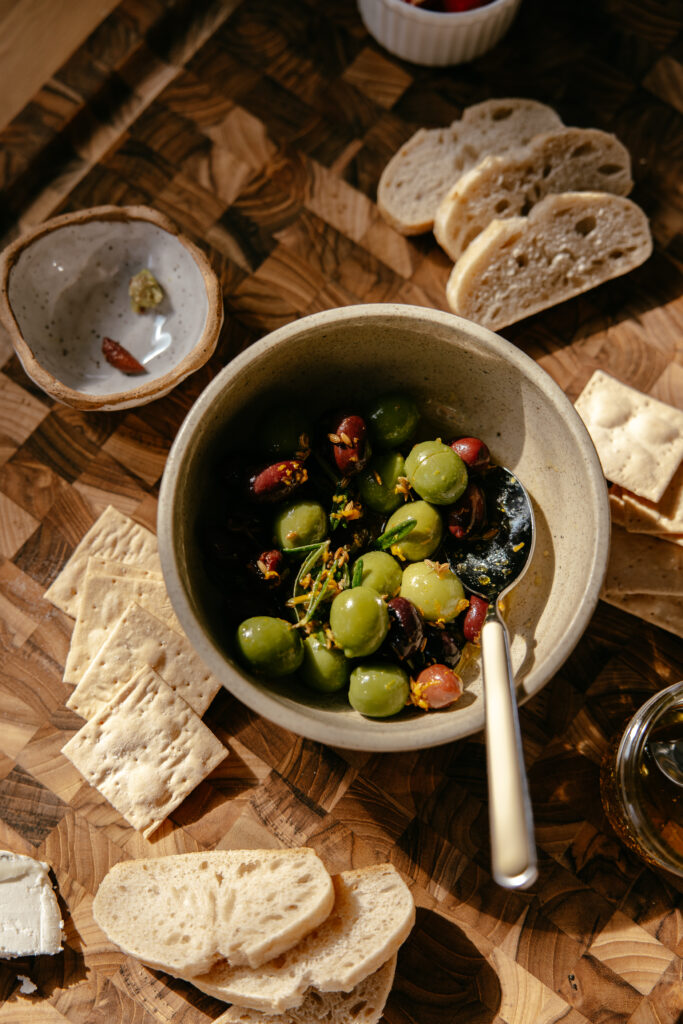 Rosemary, bay leaf, and fennel citrus-marinated olives rest in a bowl atop a wooden board.